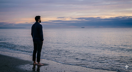 Person standing alone at the ocean's edge at dusk, looking out at an expansive quiet sky